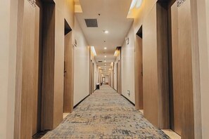 Un couloir d’hôtel avec des portes en bois, un revêtement de sol en moquette et des luminaires encastrés.