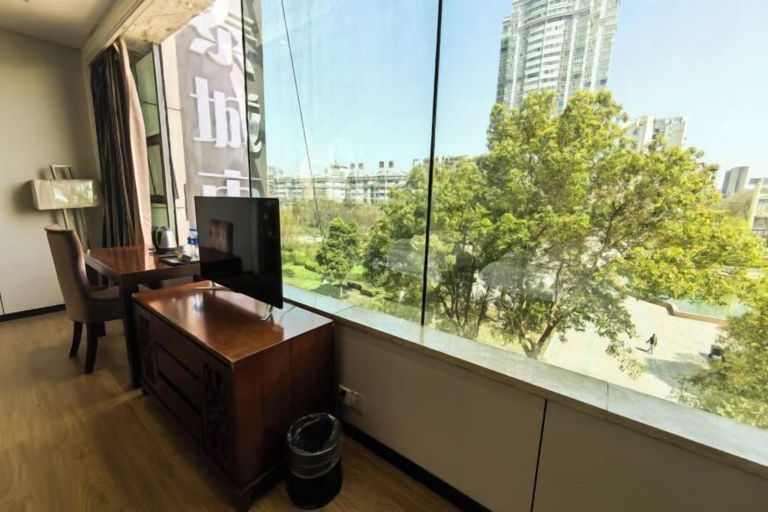 Una habitación con un ventanal grande, un escritorio de madera, un televisor y un cubo de basura.
