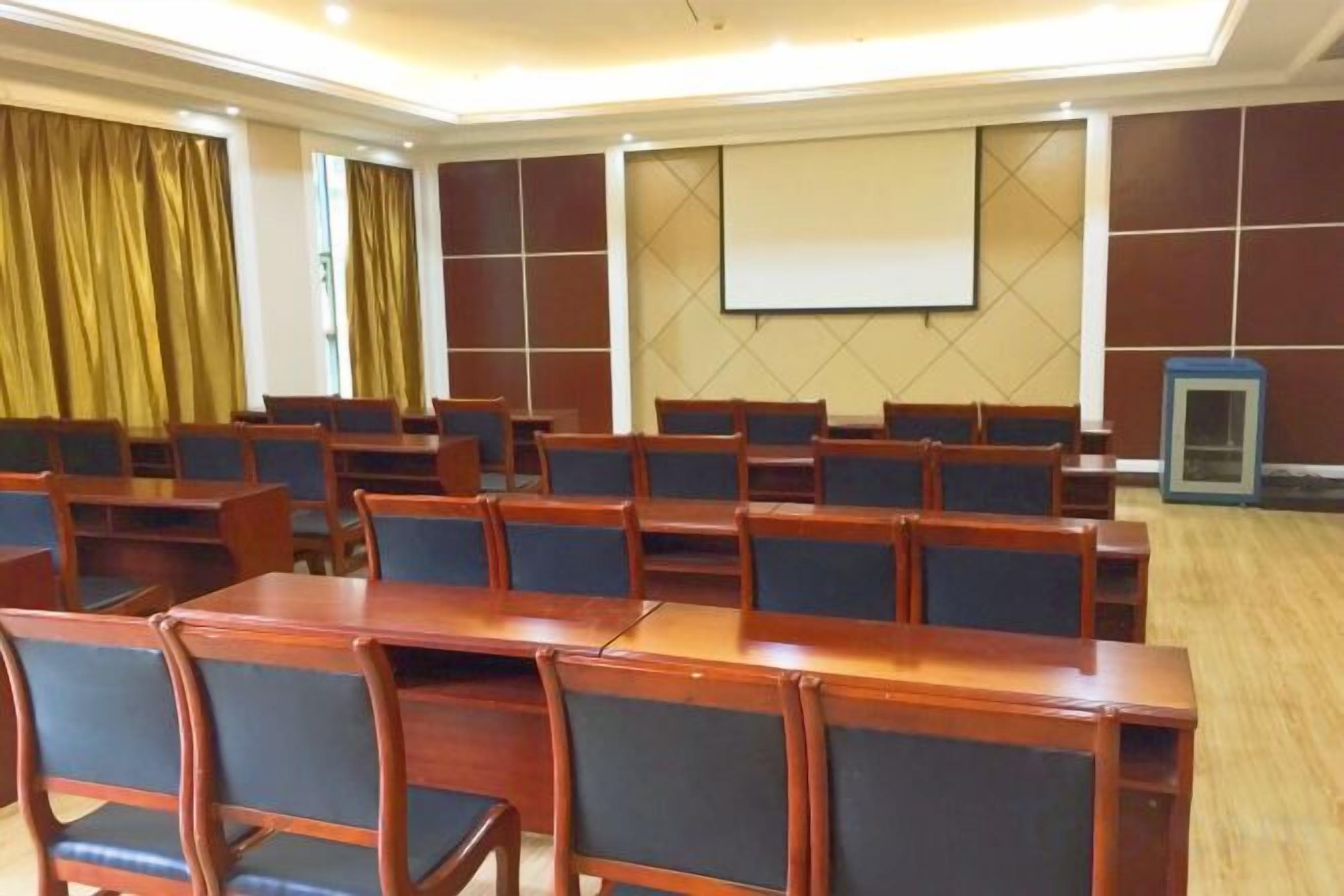 A conference room with rows of wooden tables and chairs, a projector screen, and a presentation screen.