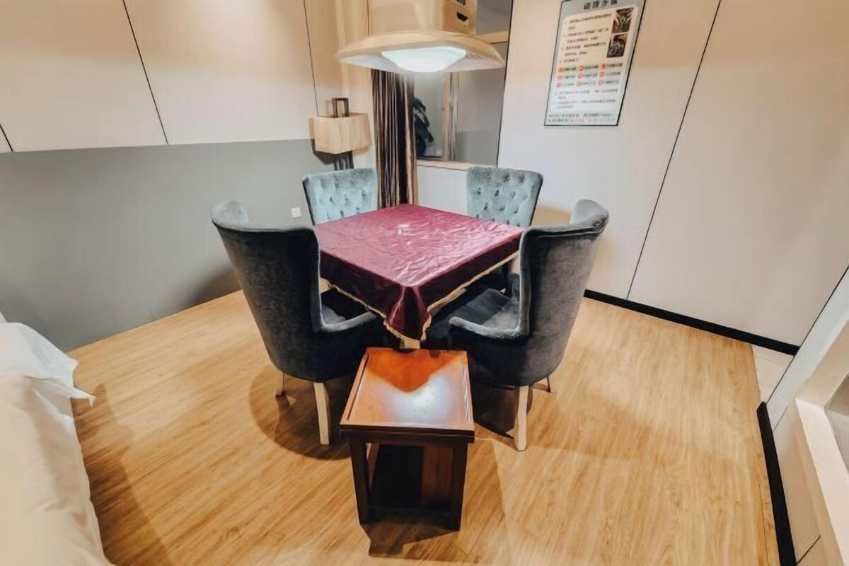 Une pièce de taille modeste avec un plancher en bois, une petite table recouverte d’une nappe rouge, quatre chaises rembourrées, une lampe et un avis encadré accroché au mur.