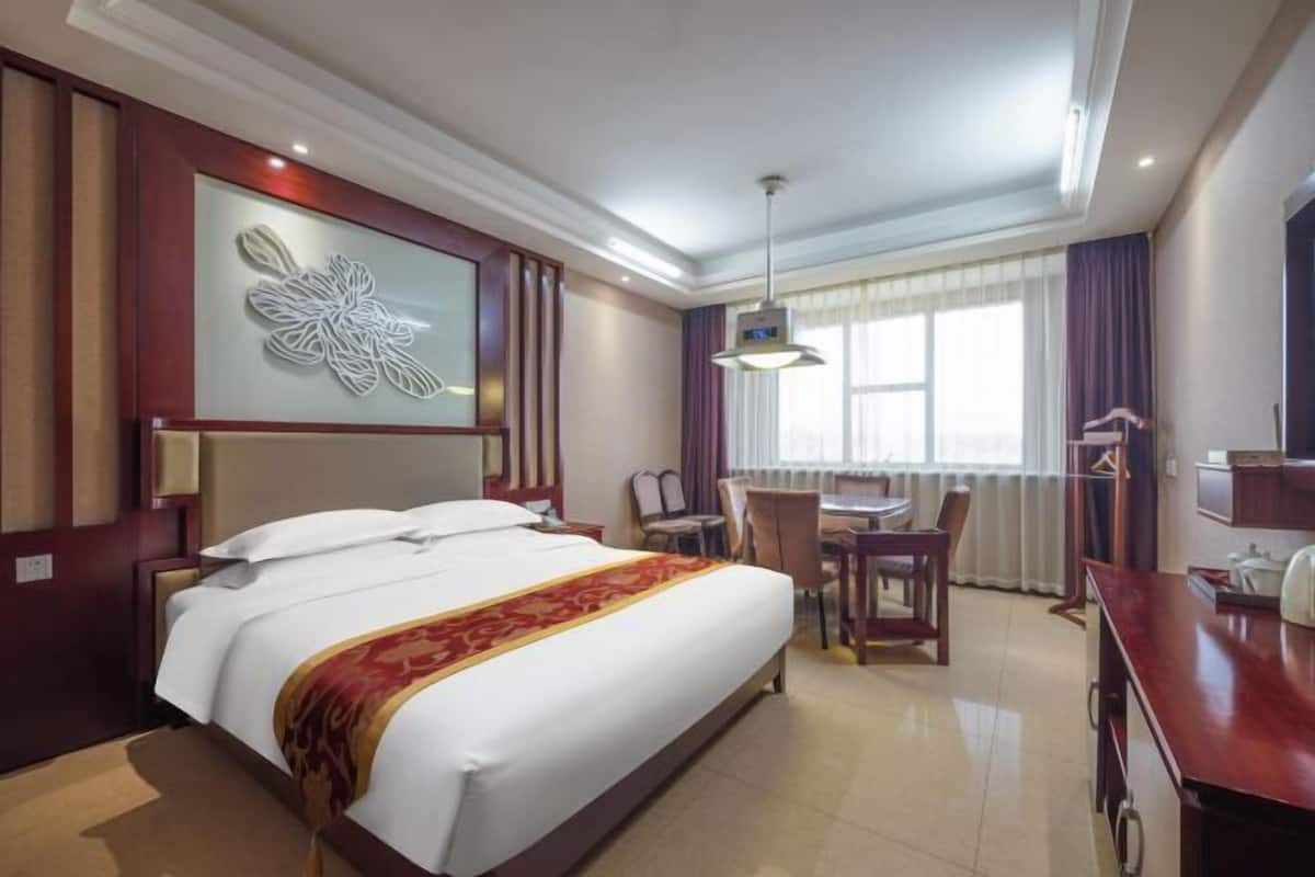 Une chambre d’hôtel avec un grand lit, un bureau avec une chaise, une table à manger et une grande fenêtre avec des rideaux.