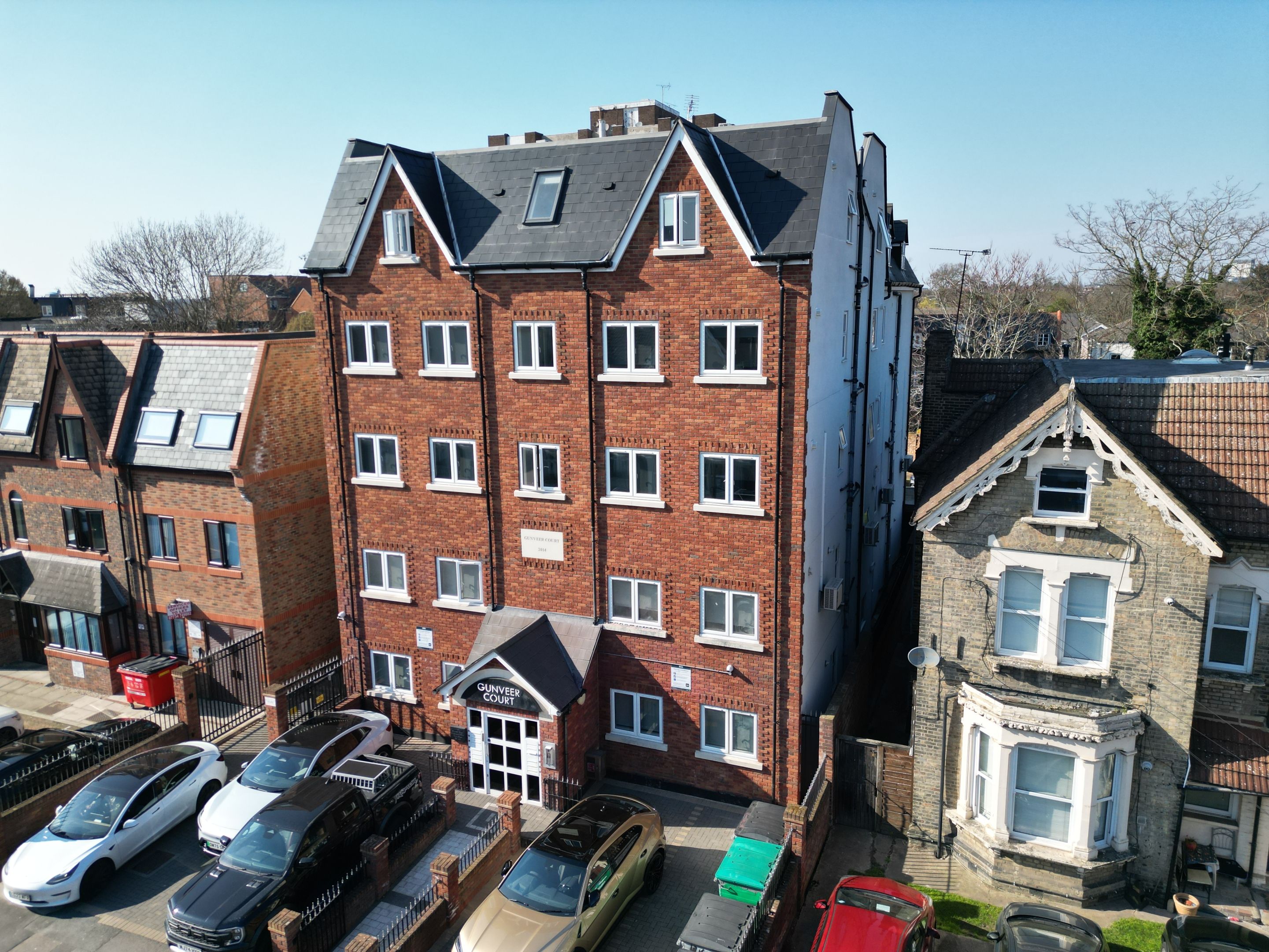 A multi-story residential building with a brick facade, multiple windows, and a parking area with cars.