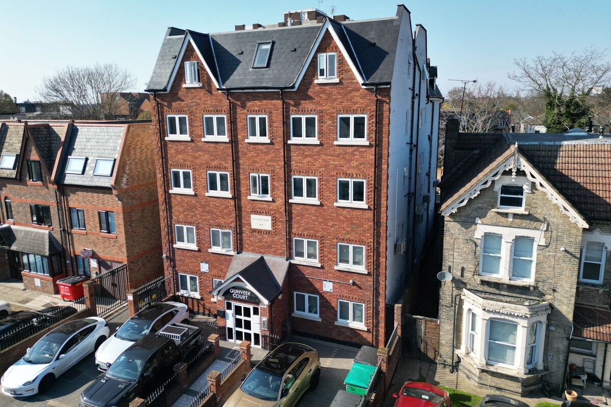 A multi-story residential building with a brick facade, multiple windows, and a parking area with cars.