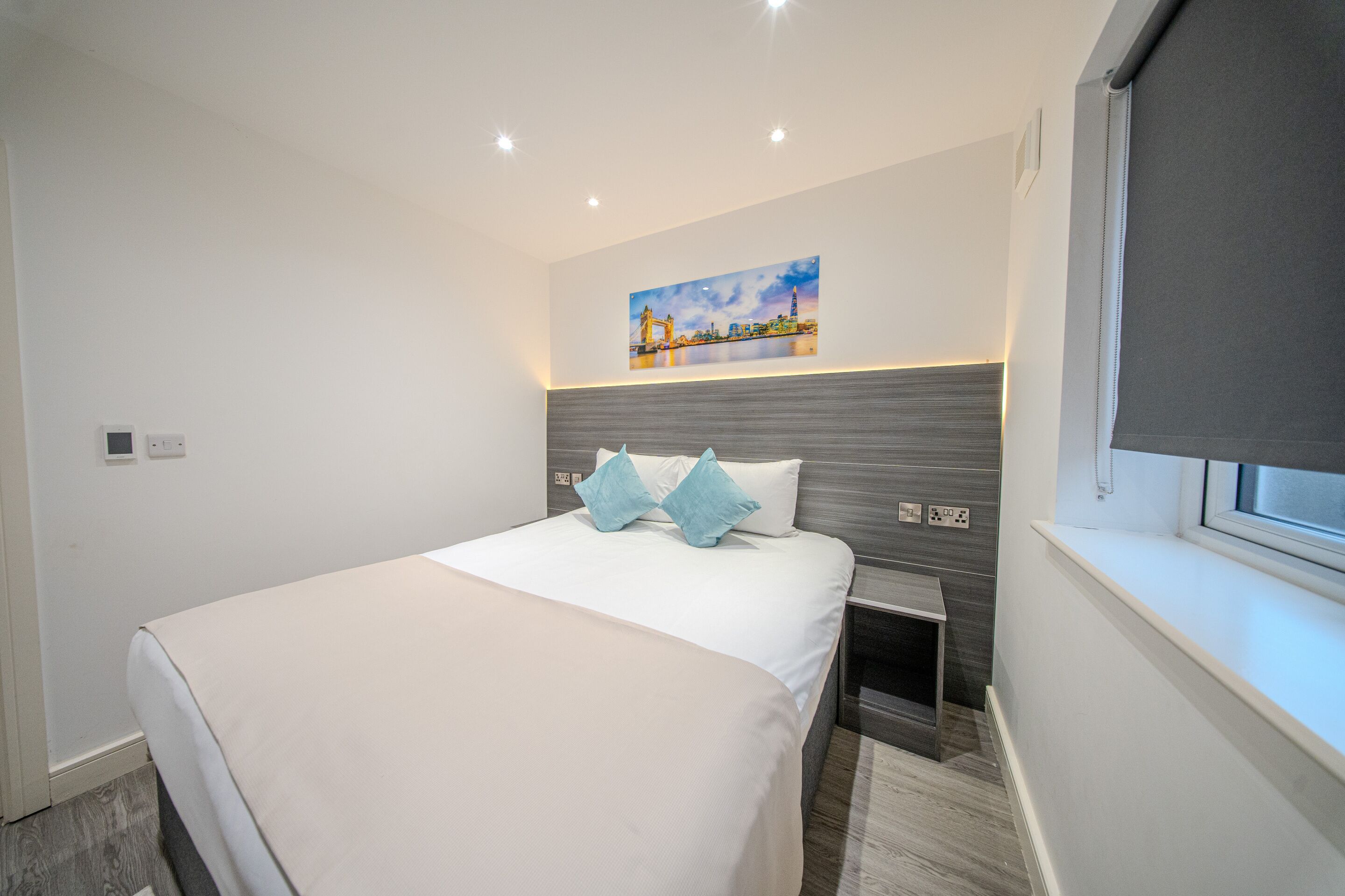 Ett modernt hotellrum med en snyggt bäddad säng, ett litet sängbord, en nattdukslampa och ett fönster med utsikt över stadsbilden.