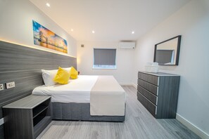 A modern hotel room with a large bed, a bedside table, a dresser, a window, and a painting of a bridge. - Ilford Apartments (Ilford)