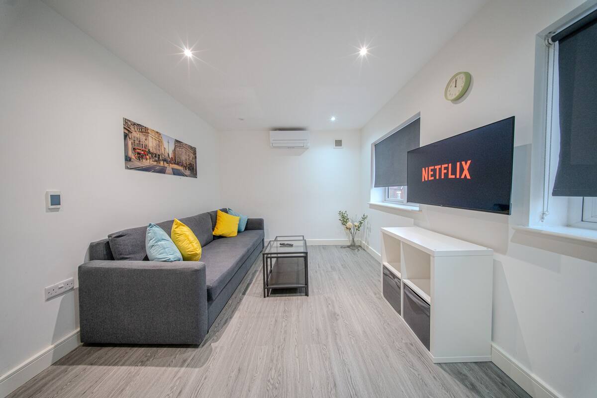 Een moderne woonkamer met een grijze bank, een salontafel, een aan de muur bevestigde televisie en een klein bijzettafeltje.