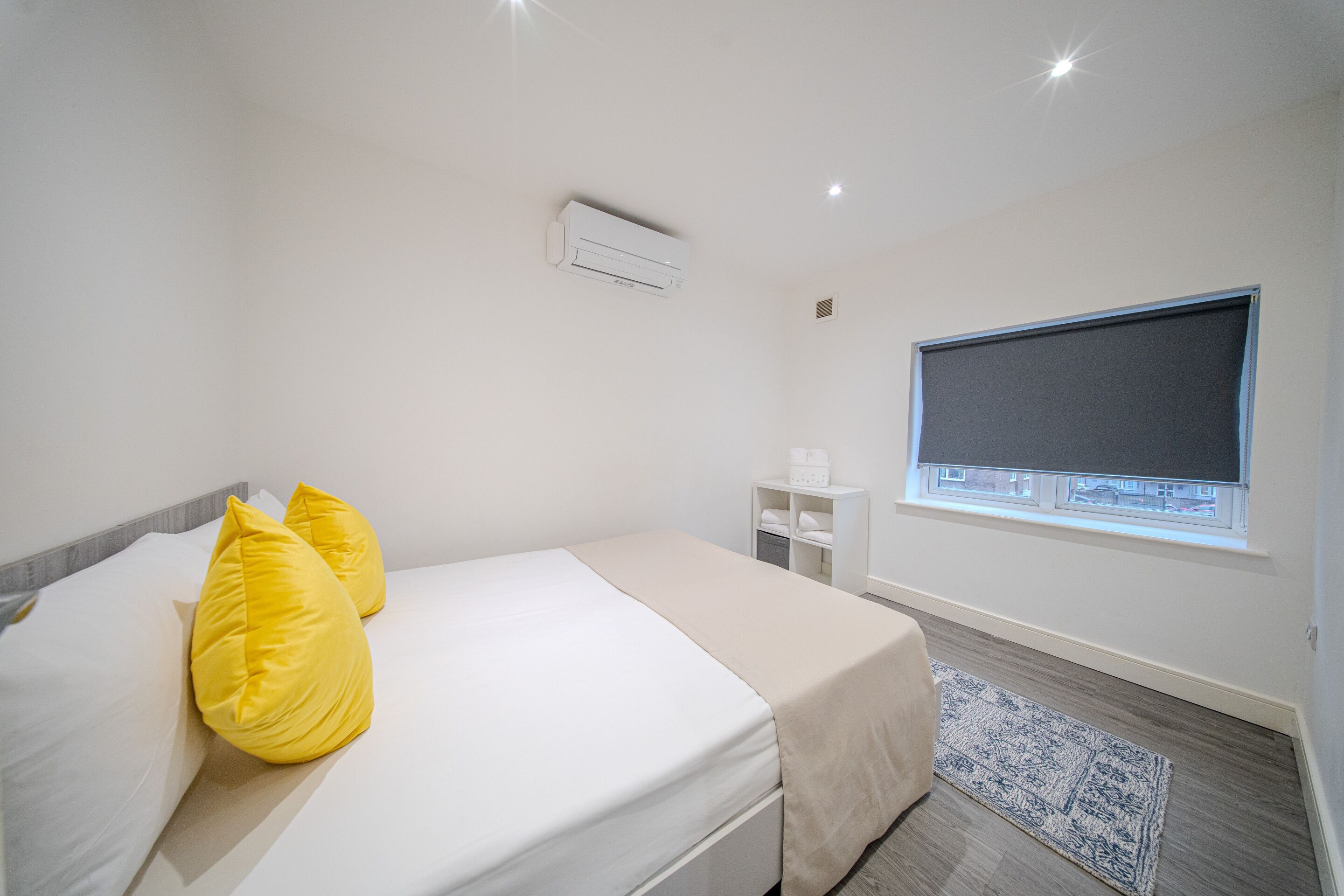 A hotel room with a bed, two yellow pillows, a window with a view, a white air conditioner, and a small bedside table with a lamp.