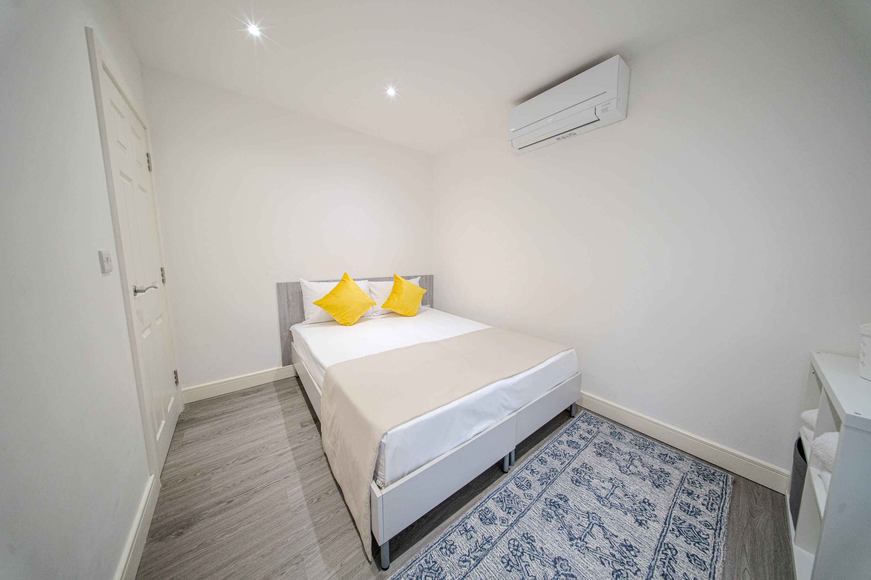 A small, clean room with a bed, a white door, a white air conditioning unit, and a patterned rug.