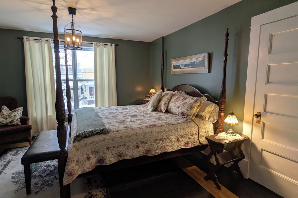 Un dormitorio con una cama con dosel, una silla, una mesita de noche con lámpara, una puerta y una ventana con cortinas.