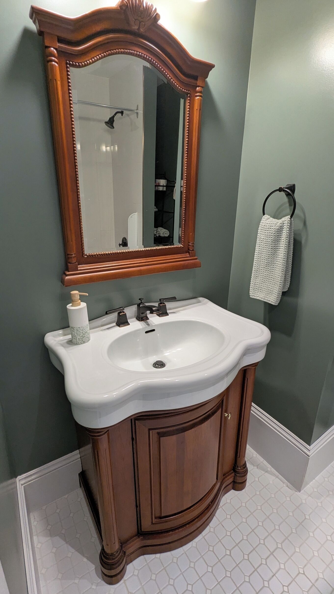 Un baño con tocador de madera, un lavabo blanco, un espejo con marco de madera y un perchero con una toalla blanca.