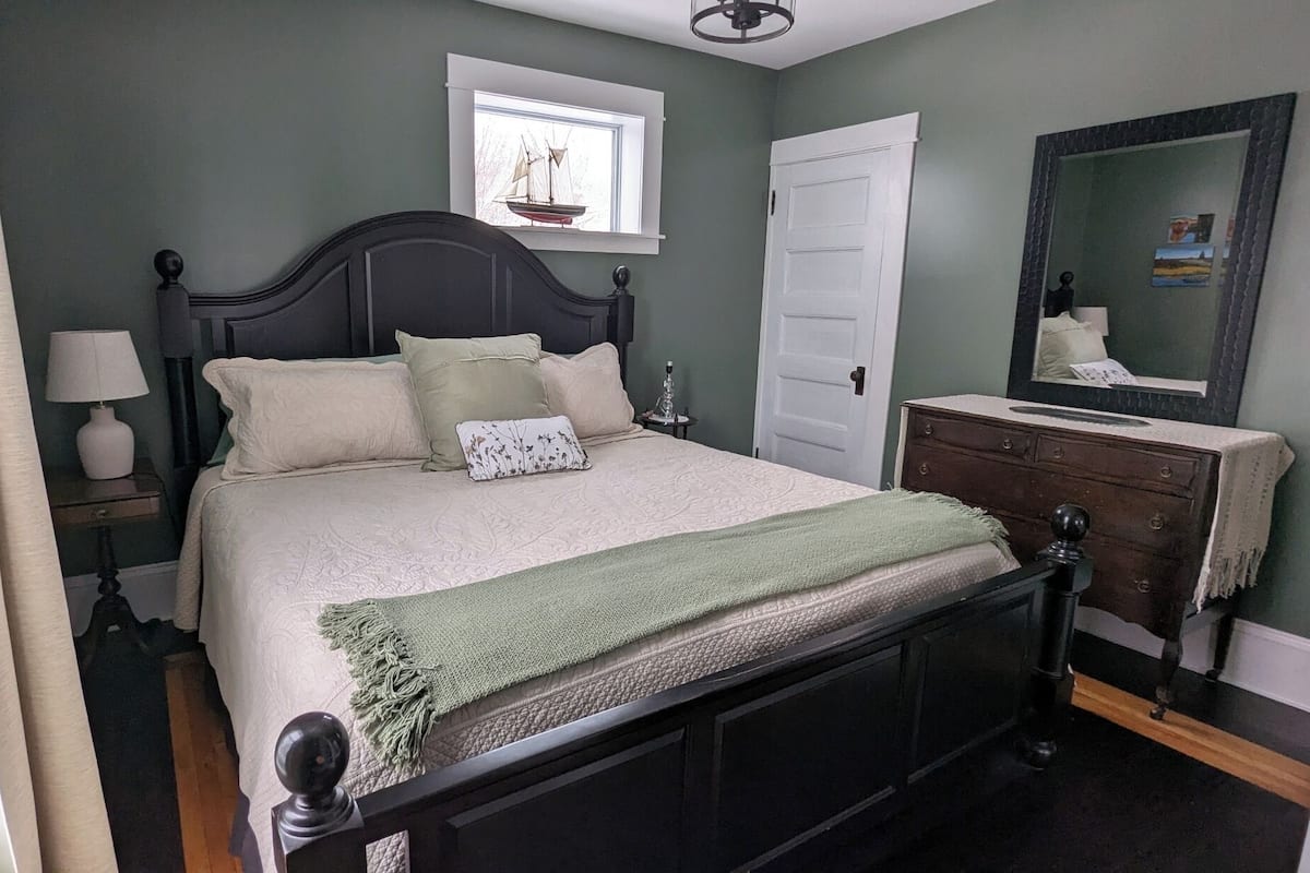 Un dormitorio con cama, mesita de noche, cómoda, espejo y ventana decorada con un velero.
