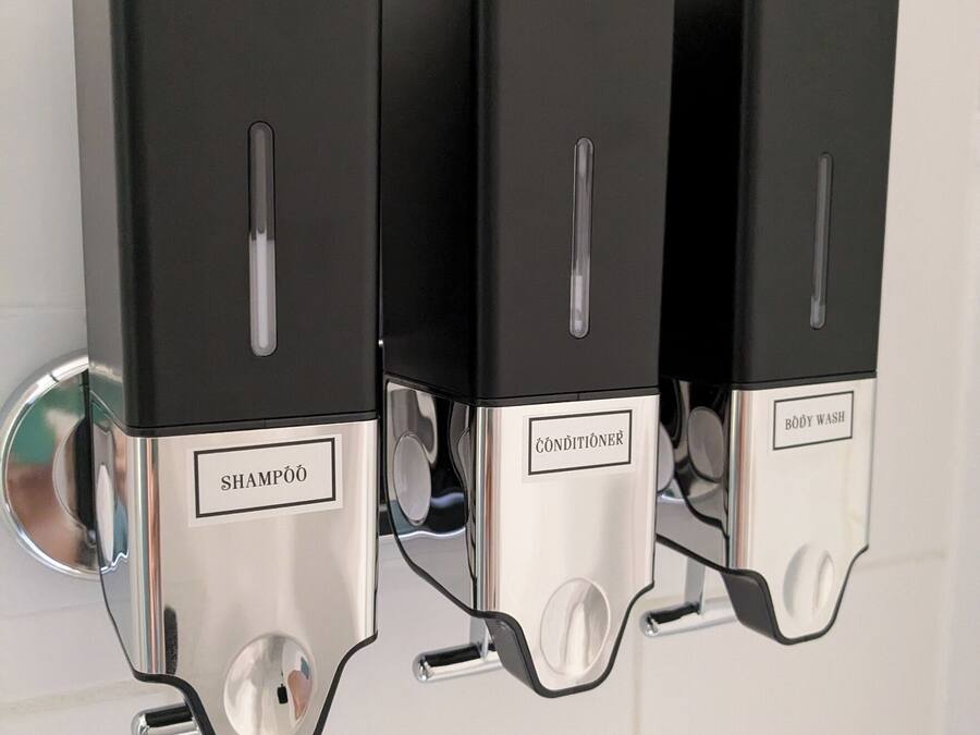 A wall-mounted dispenser with three compartments labeled 'SHAMPOO,' 'CONDITIONSER,' and 'BODY WASH.'