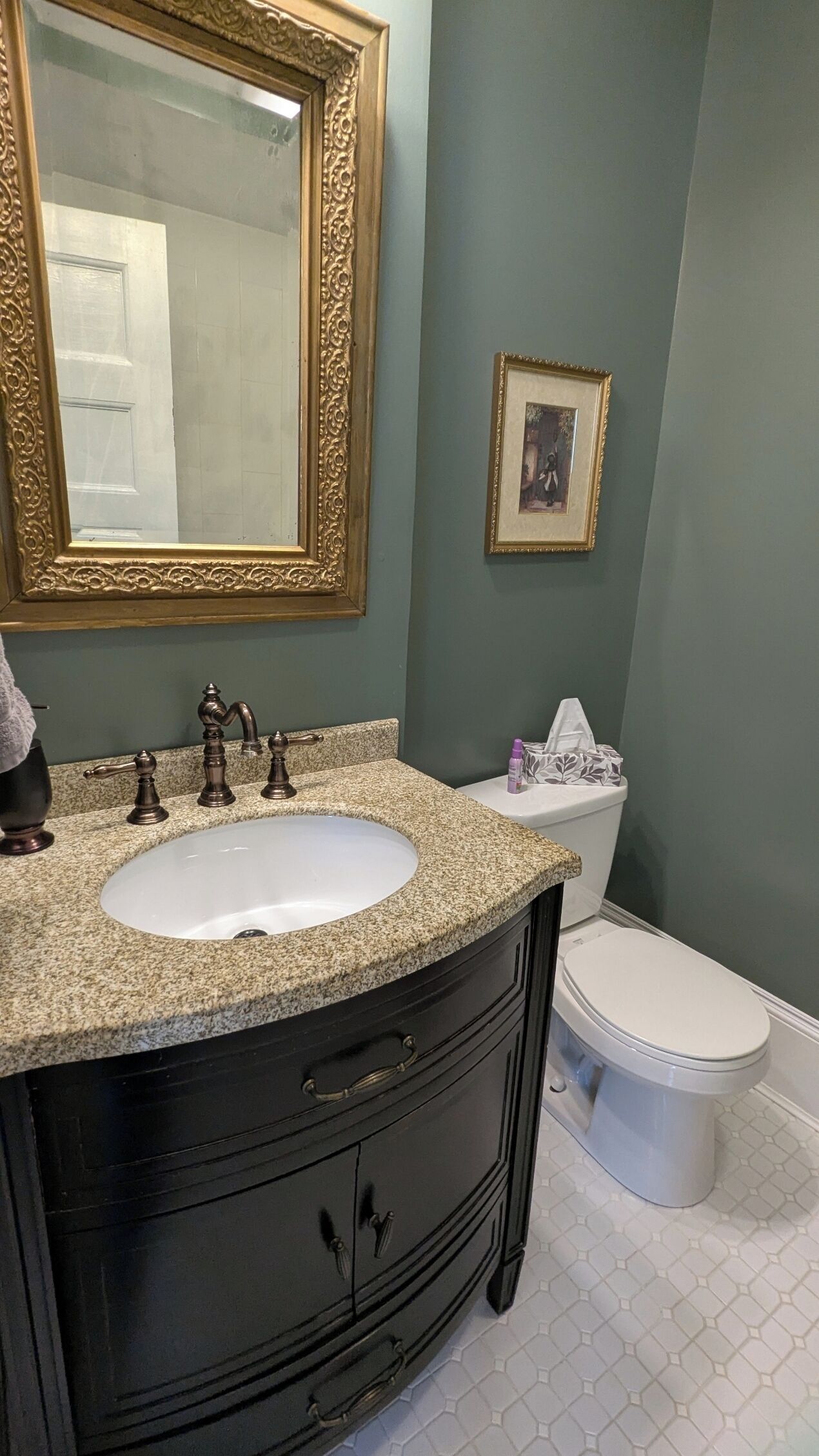 A bathroom with a granite countertop sink, a black vanity, a gold-framed mirror, a white toilet, and a framed picture on the wall.