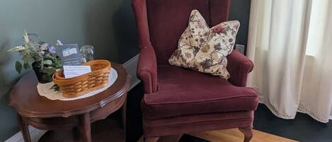 Una habitación de esquina con una pared verde oscuro, un sillón rojo con un cojín floral, una mesita redonda con una cesta y un cuadro enmarcado, y una alfombra estampada.