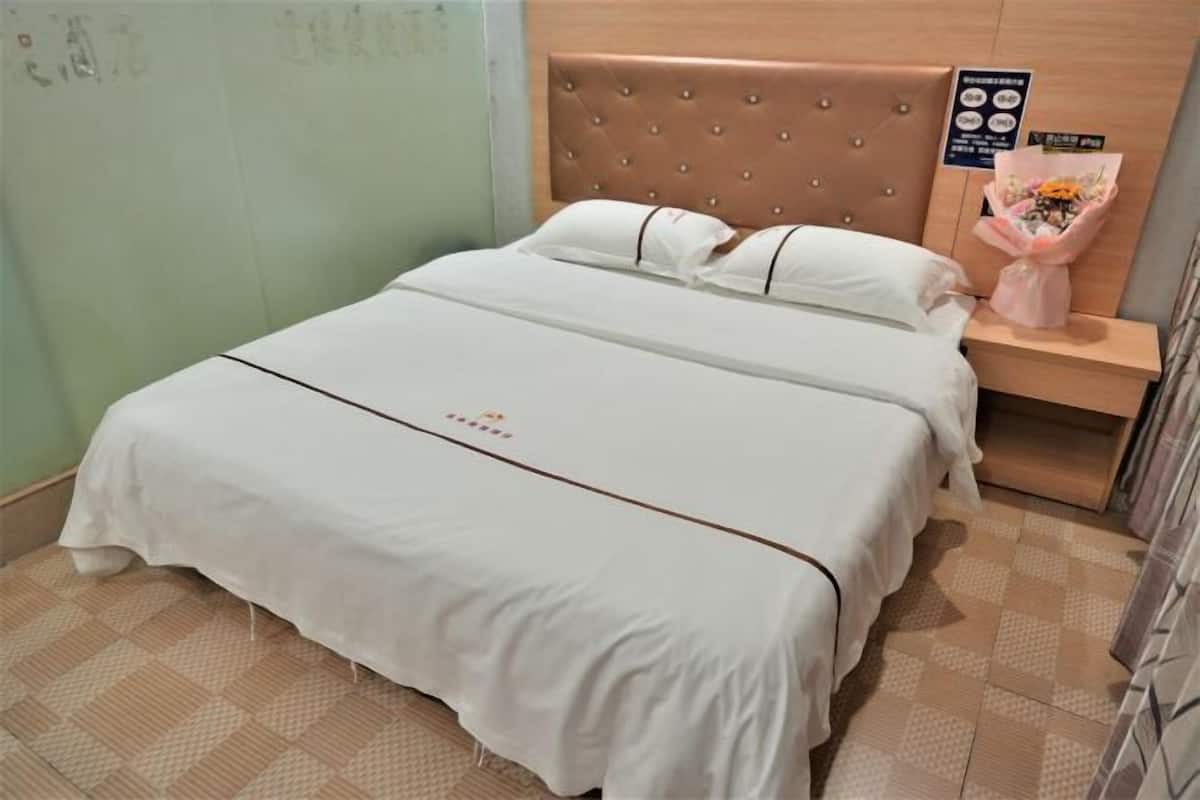 Cama bem arrumada com colcha branca e cabeceira marrom. Um criado-mudo exibe um buquê de flores e uma pequena placa. O quarto possui um padrão de piso quadriculado.