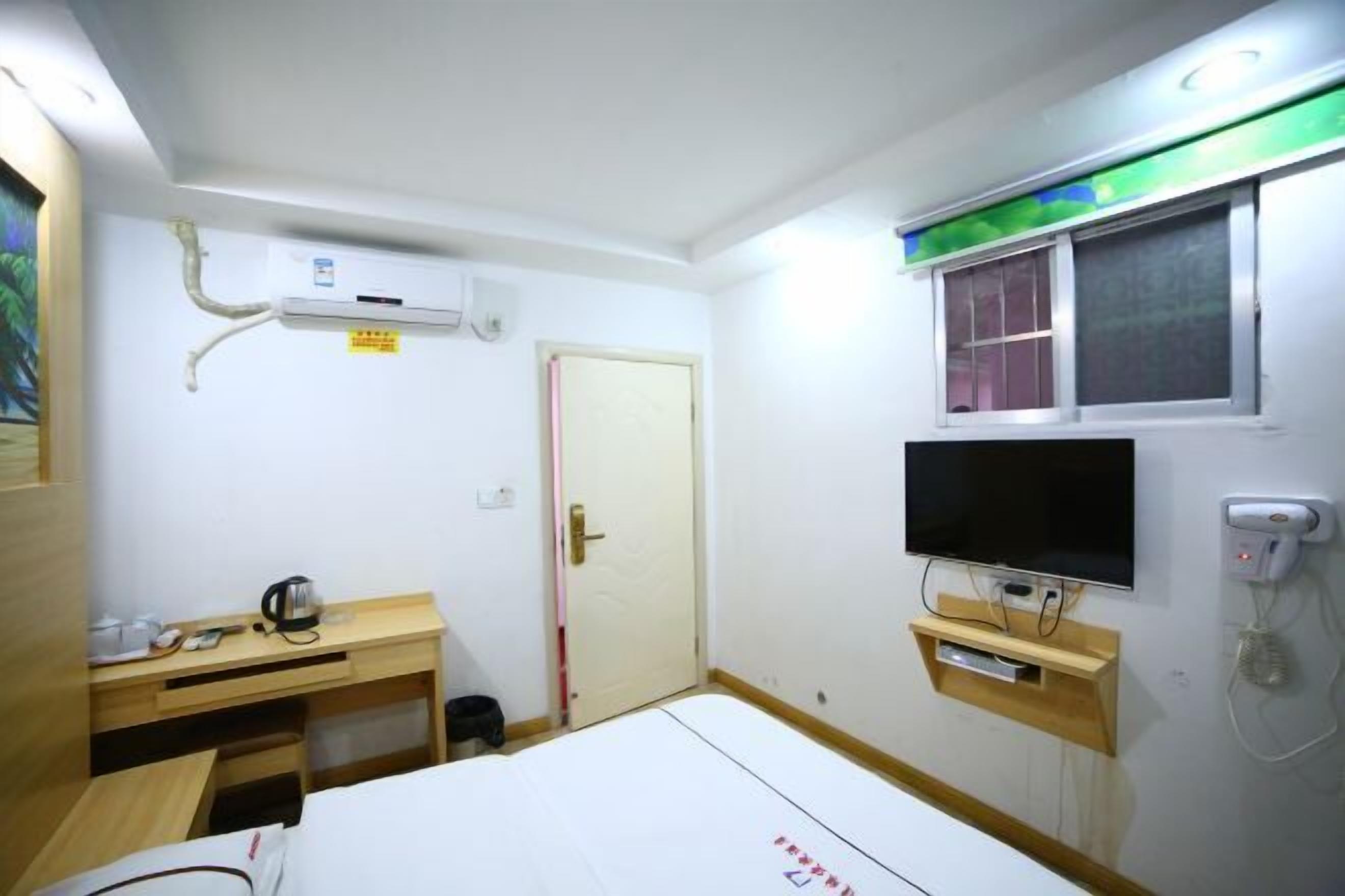 Een hotelkamer met een bed, een bureau, een televisie, een airconditioning en een raam met luxaflex.