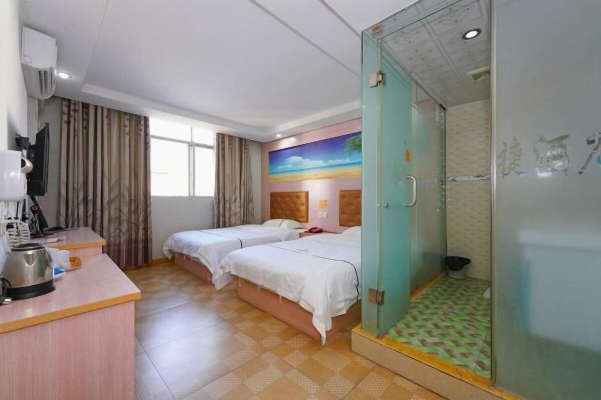 A hotel room with two beds, a desk with a kettle, a TV, and a shower area.
