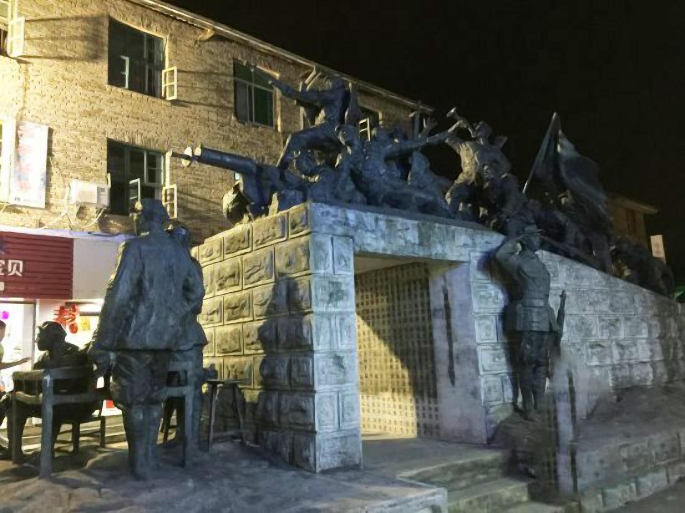 A stone monument with figures, located in front of a building with a visible sign.