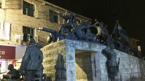 Een stenen monument met figuren in verschillende houdingen, voor een gebouw met een zichtbare winkelgevel.