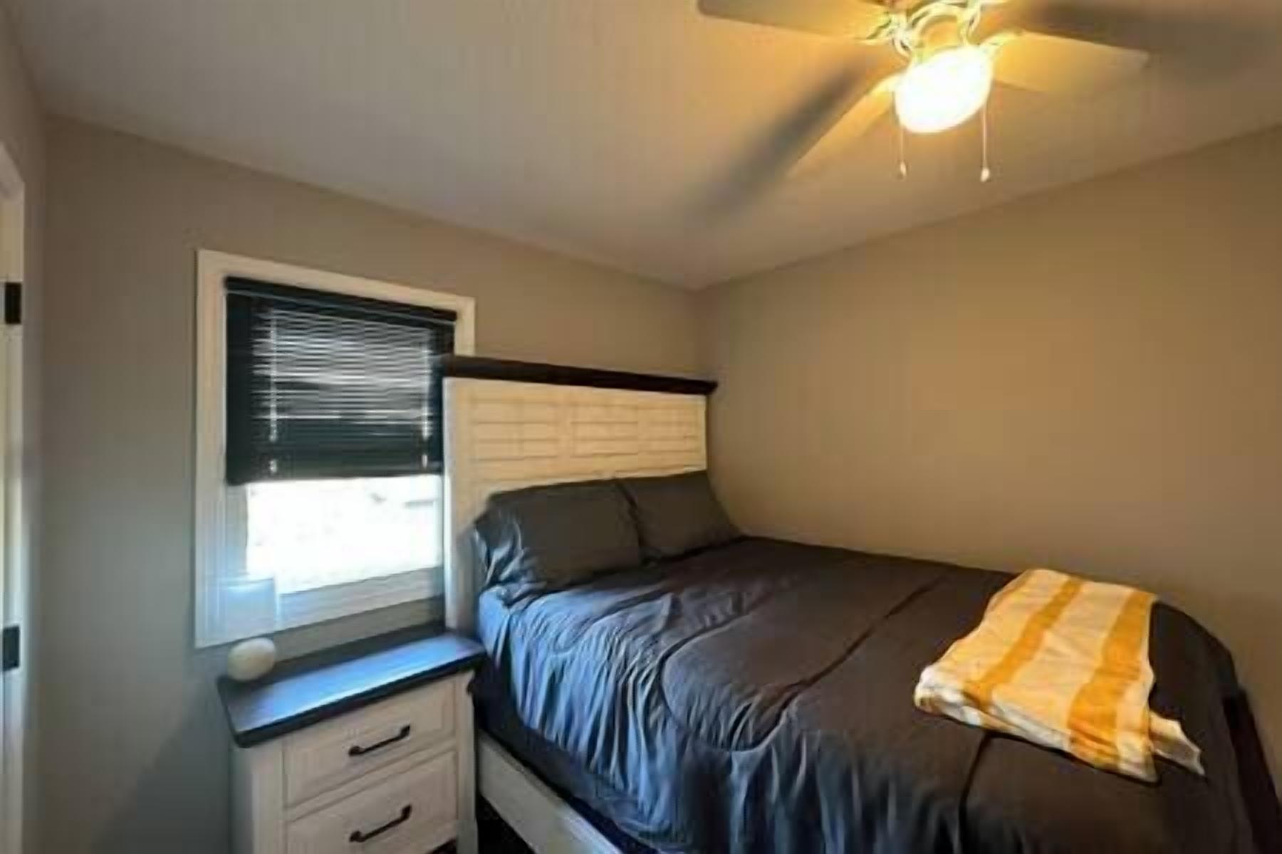 A bedroom with a bed, a nightstand, a window with blinds, and a ceiling fan.