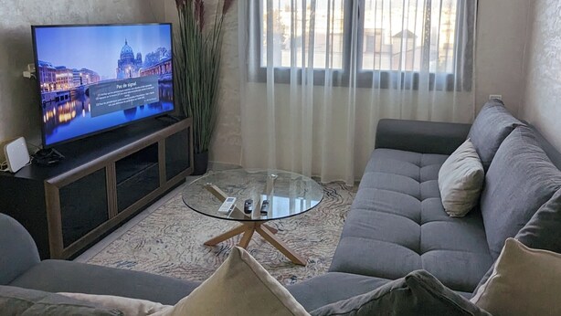 A living room with a modern chandelier, a flat-screen TV, a sofa set, and a large window with sheer curtains. - Fez apartment (Fès)