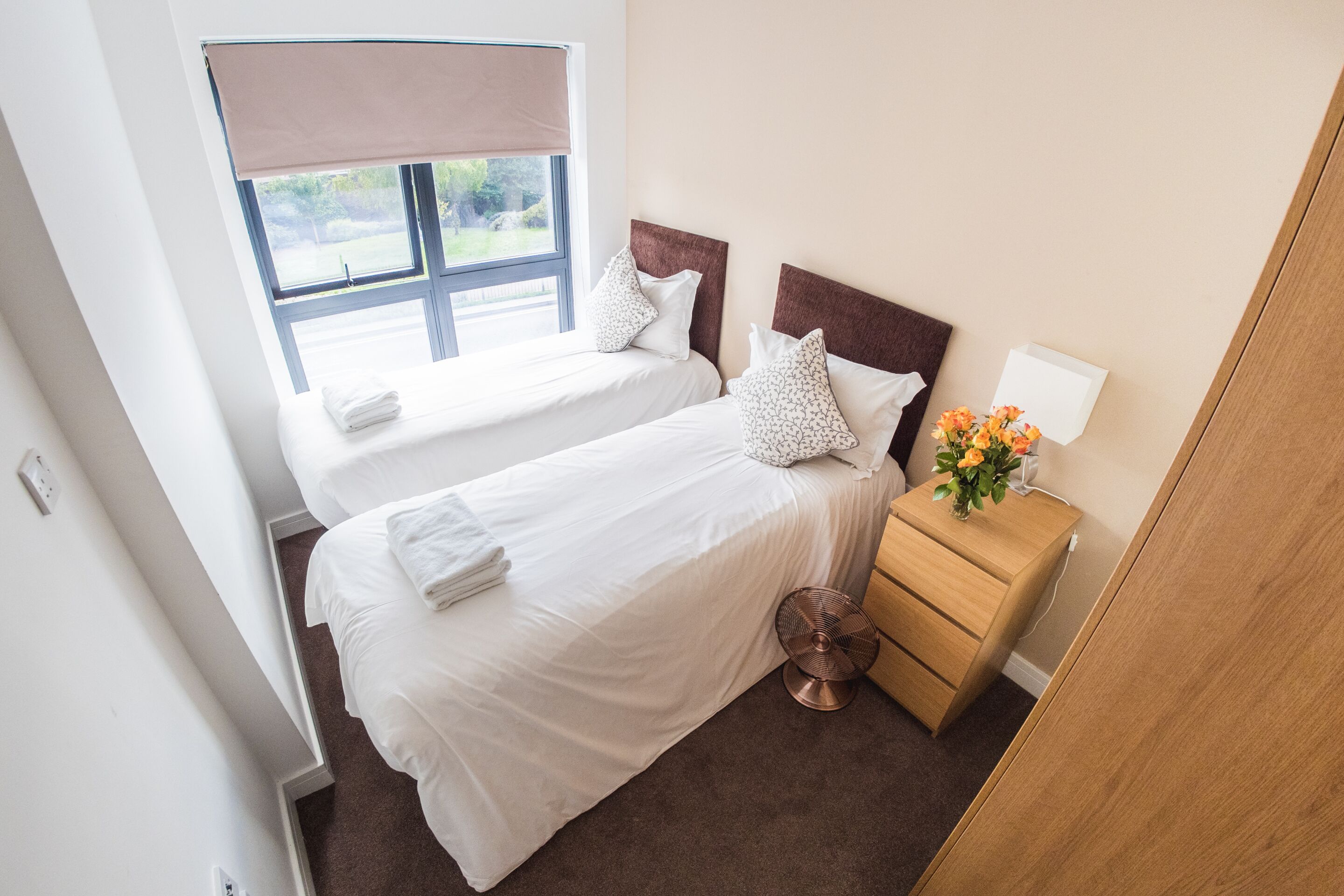 A hotel room with two beds, a wooden nightstand with a fan, a window with a blind, and a small table with a vase of flowers.