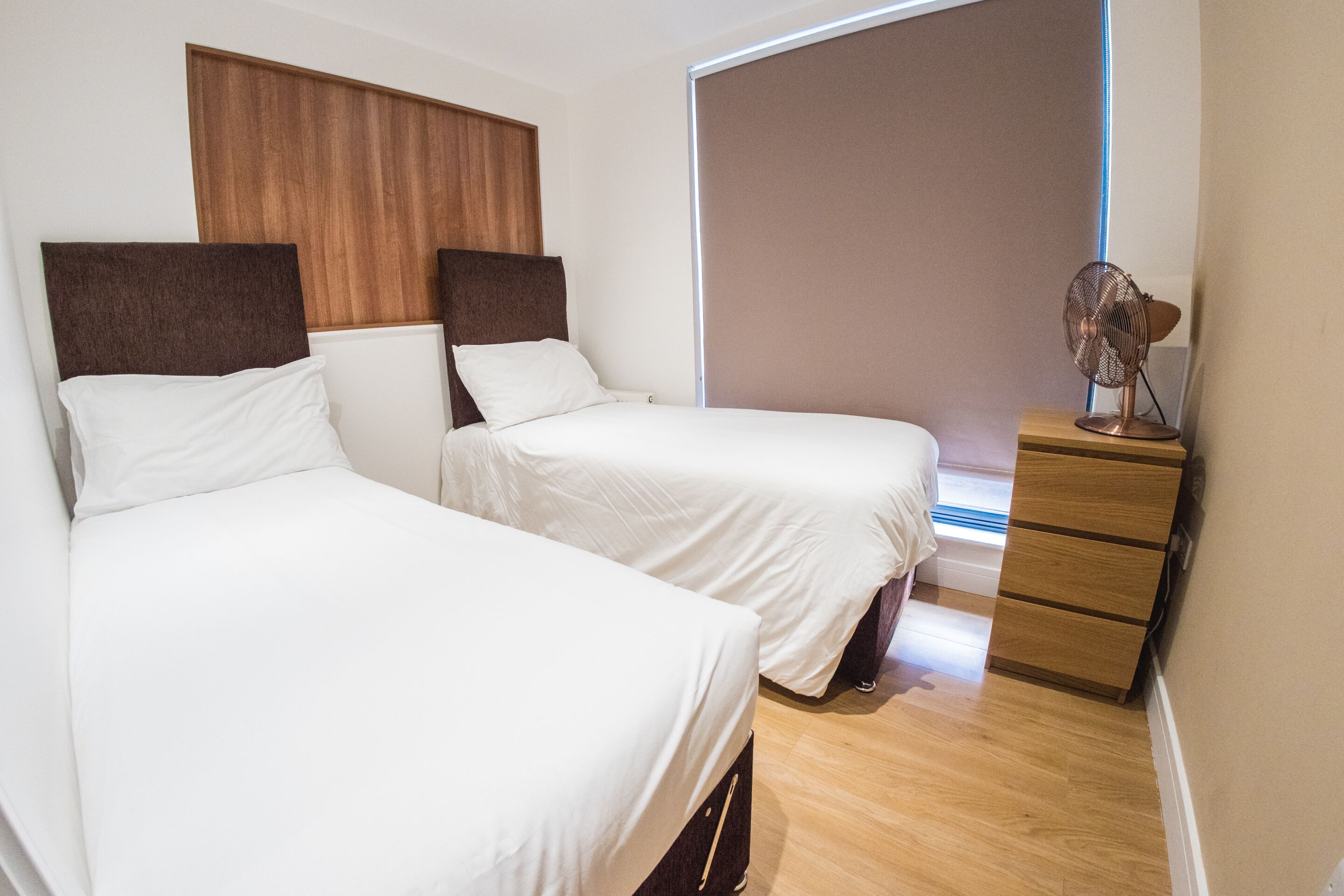 Una habitación de hotel con dos camas, un cabecero y un aparador de madera, y un ventilador de pie.