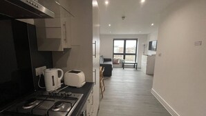 A modern kitchen with a gas stove, a kettle, and a toaster. There is a dining area with a table and chairs, and a large window providing a view of the outside. - Ilford Tower Apartments (Ilford)