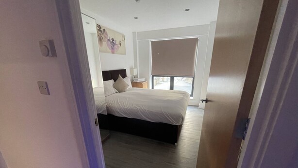 A bedroom with a bed, a nightstand, a window, and a door. - Ilford Tower Apartments (Ilford)