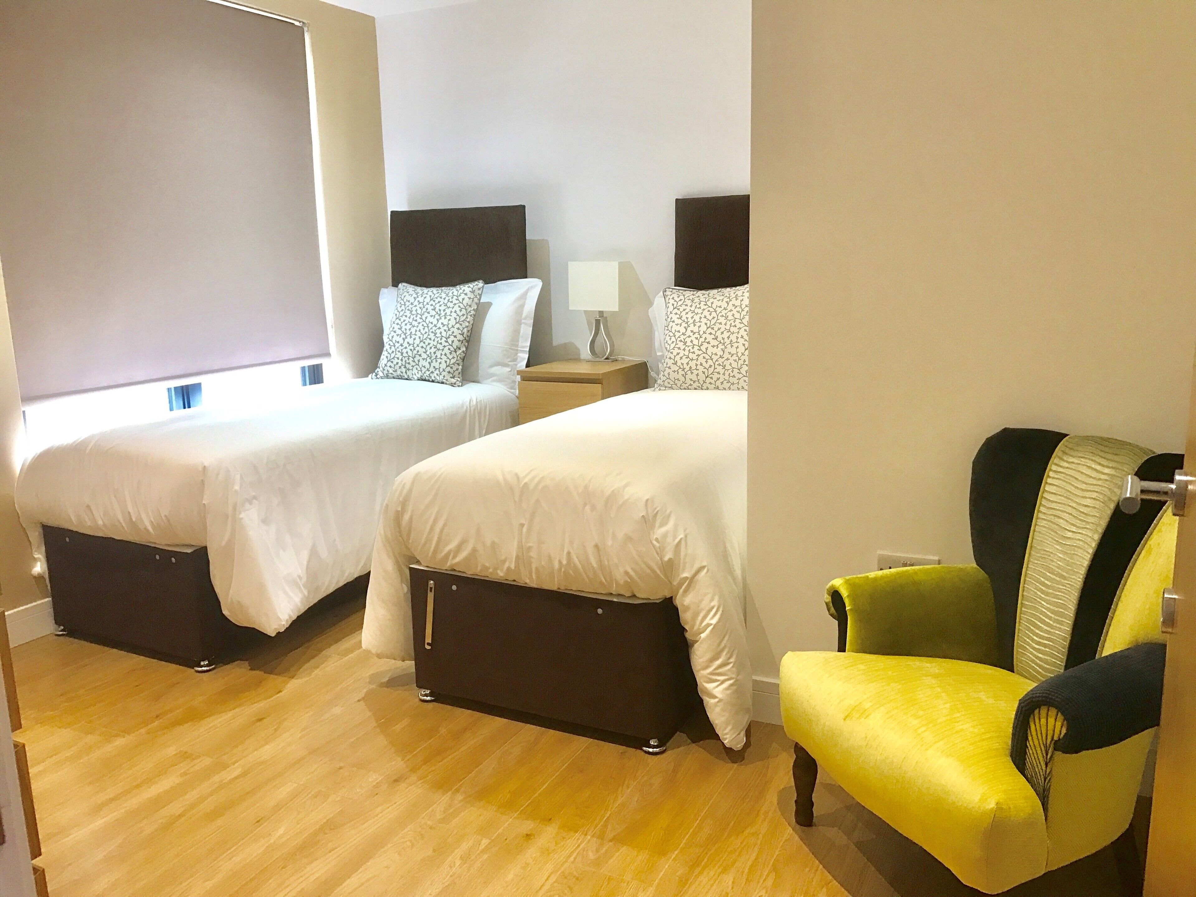 A hotel room with two beds, a nightstand, a lamp, and a yellow armchair.