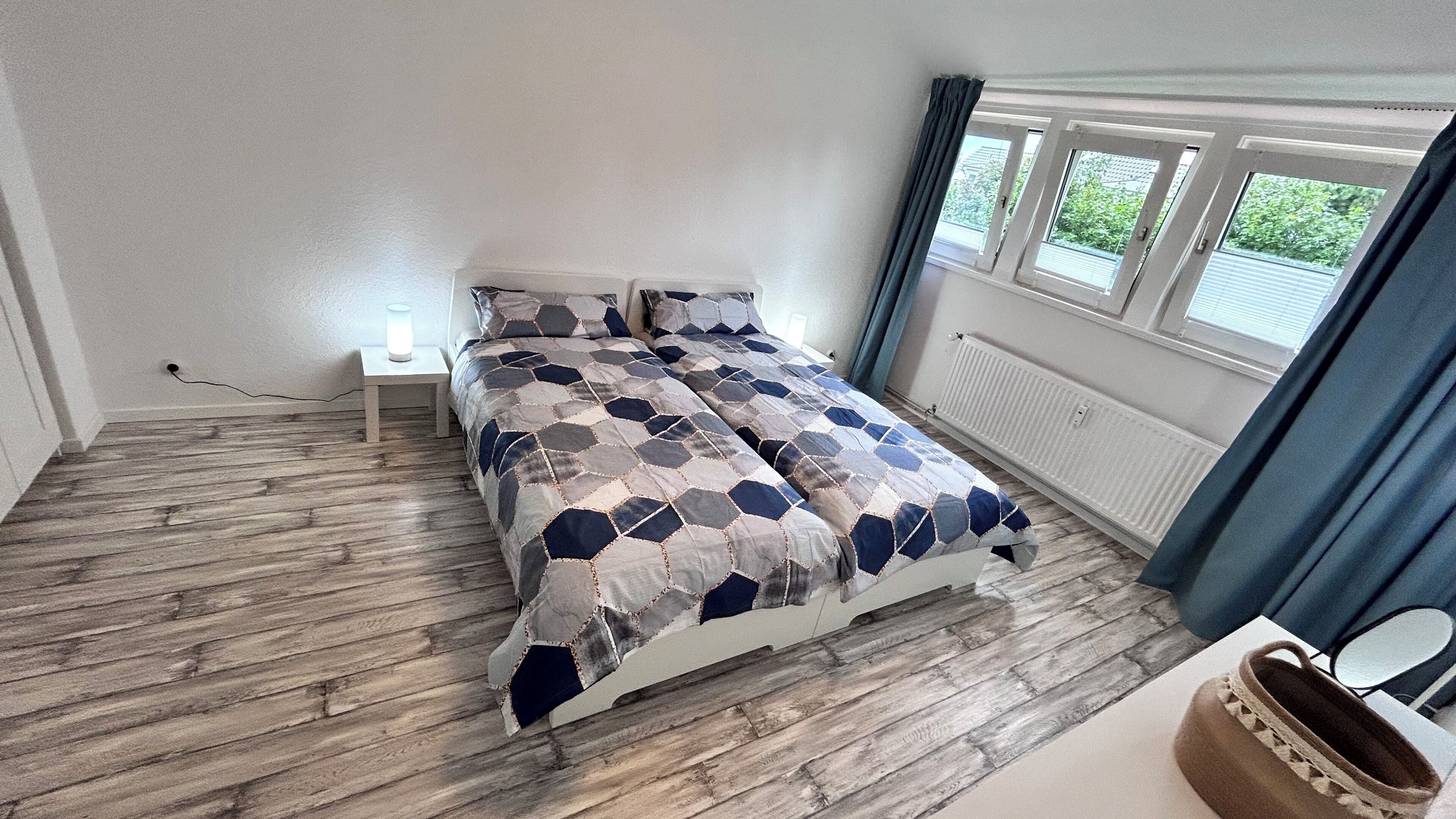 A bedroom with a bed, a nightstand, a lamp, a window with curtains, and a radiator.