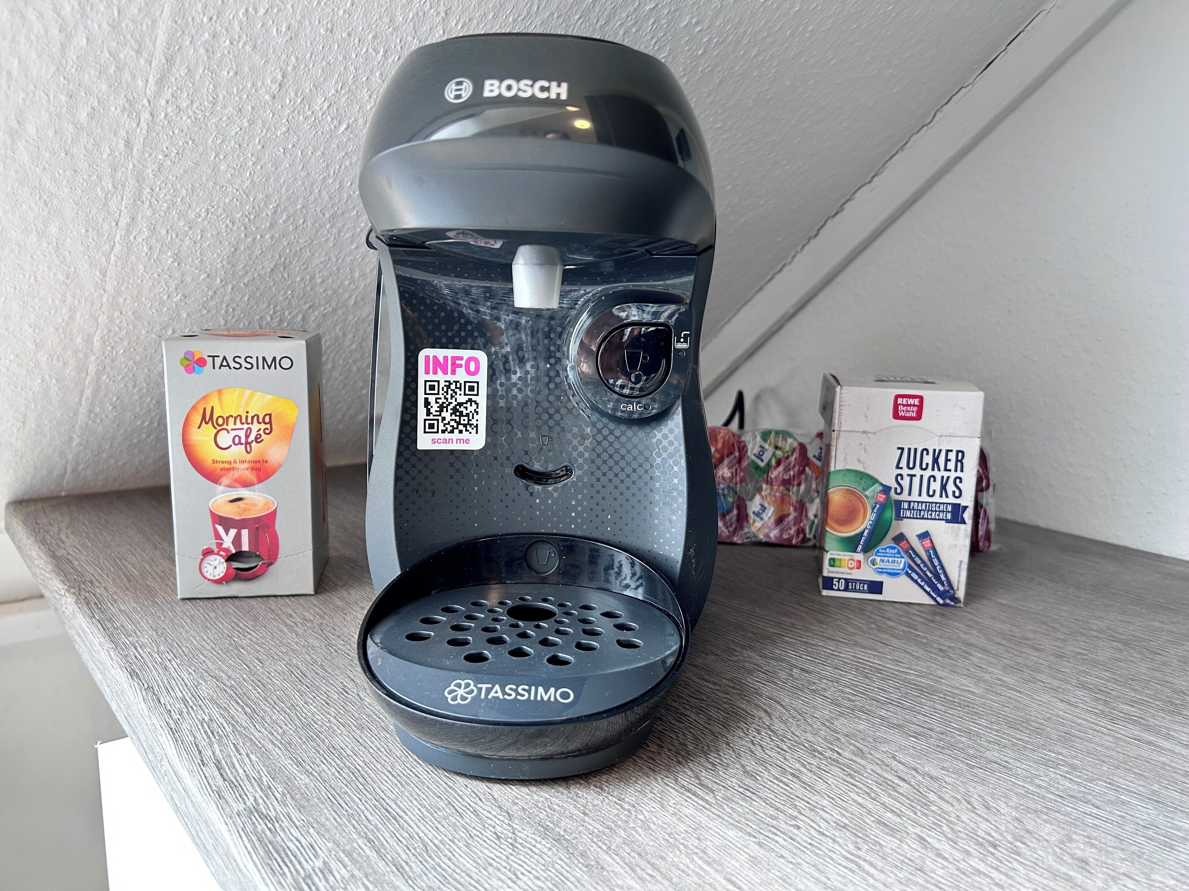Une machine à café Tassimo sur une étagère, avec une boîte de dosettes Tassimo et une boîte de bâtonnets de sucre.