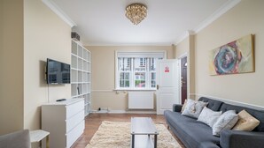 A modern living room with a grey sofa, a white entertainment unit, a flat-screen TV, and a large window with a view of a building outside. - Elegant 3Bedroom Retreat (London)