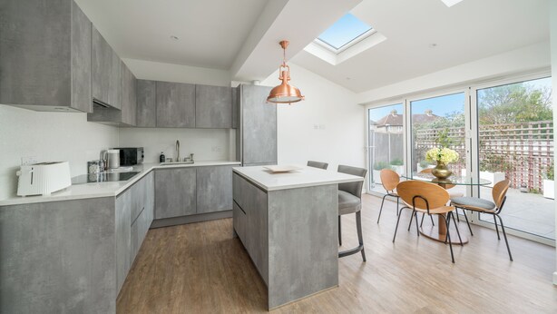 A modern kitchen with grey cabinetry, a central island, and a dining area with a round table and chairs. - Elegant 3Bedroom Retreat (London)