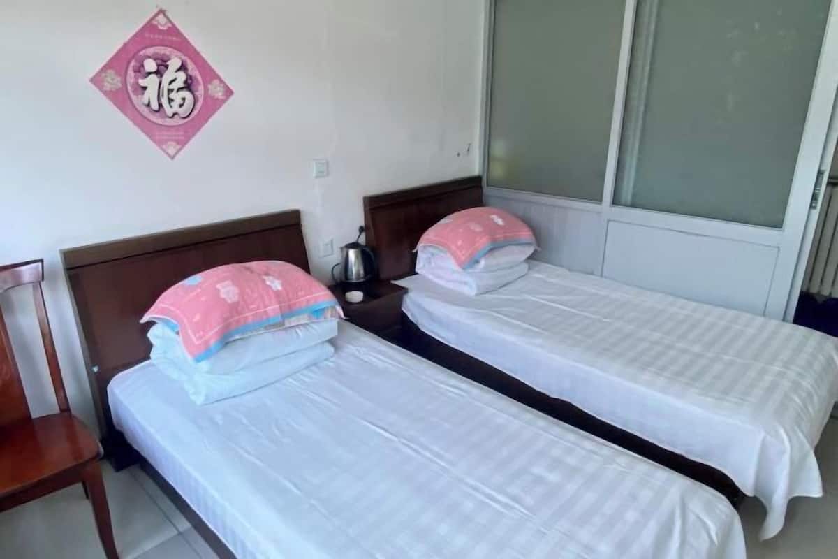Quarto com duas camas, uma cadeira de madeira, uma mesa de cabeceira com chaleira e um objeto decorativo na parede.