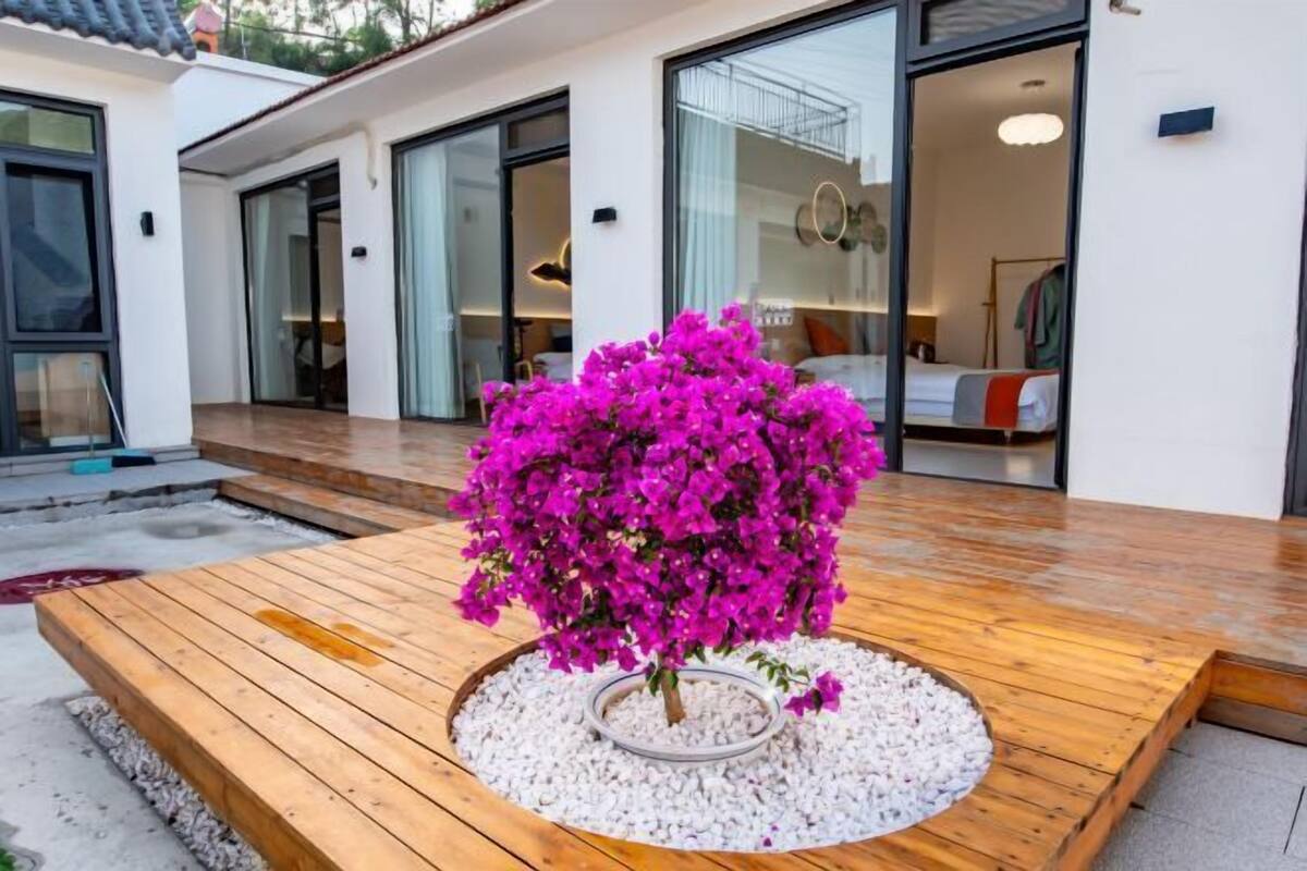 Edifício moderno com deck de madeira, planta de bougainvilleira em vaso e quarto visível com cama e mesa de cabeceira.