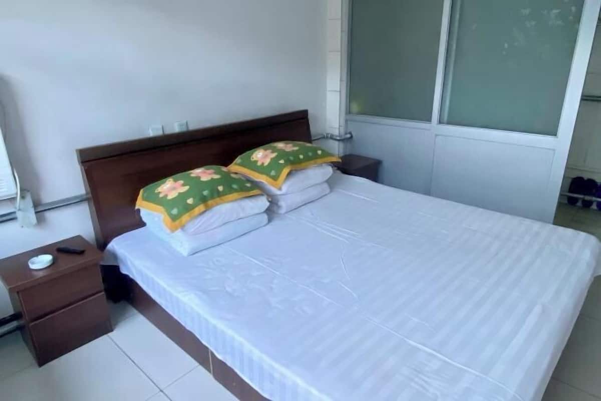 Uma cama bem arrumada com almofadas, um criado-mudo de madeira com controle remoto e uma janela com vidro fosco.