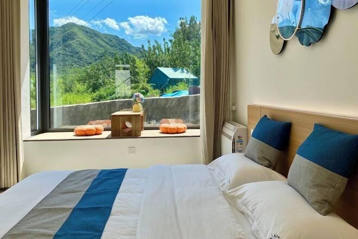 Quarto de hotel com uma cama grande, vista para montanhas, uma mesinha com abajur e uma janela com cortinas.