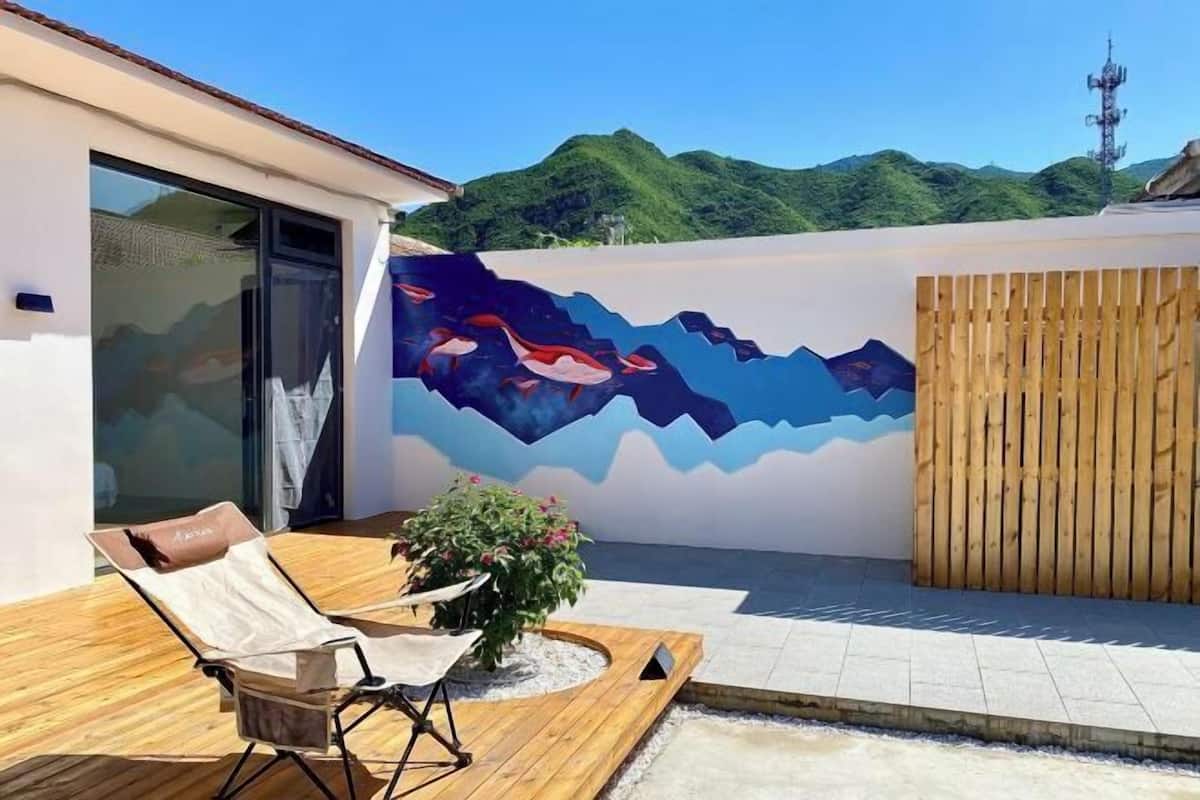 Uma área de estar moderna ao ar livre, com um mural, cerca de madeira e vista para colinas verdes.