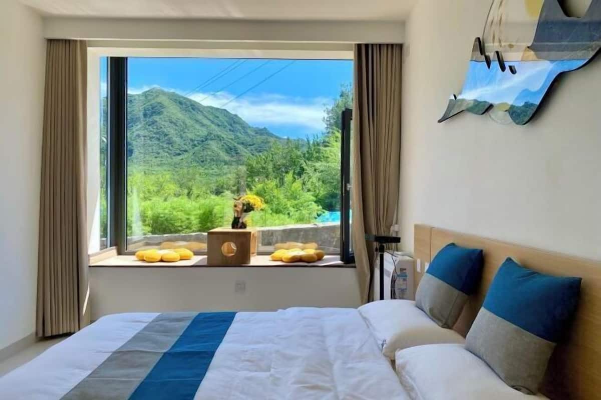 Quarto de hotel com uma janela ampla, cama com roupa de cama branca e azul, e vista para montanhas e vegetação.