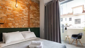 A hotel room with a brick wall, a bed with white linens, a grey headboard, a grey curtain, a small round table with a chair, and a view of the outside. - Zest Teodoro (São Paulo)