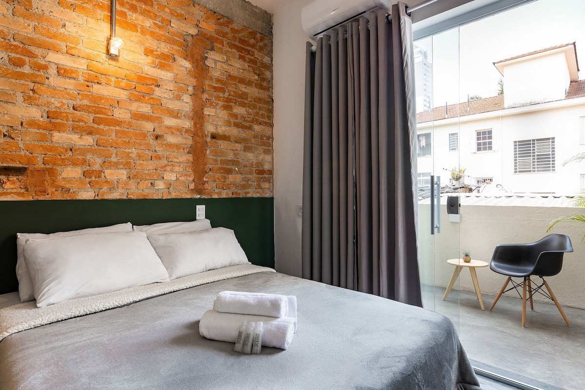 A hotel room with a brick wall, a bed with white linens, a grey headboard, a grey curtain, a small round table with a chair, and a view of the outside.