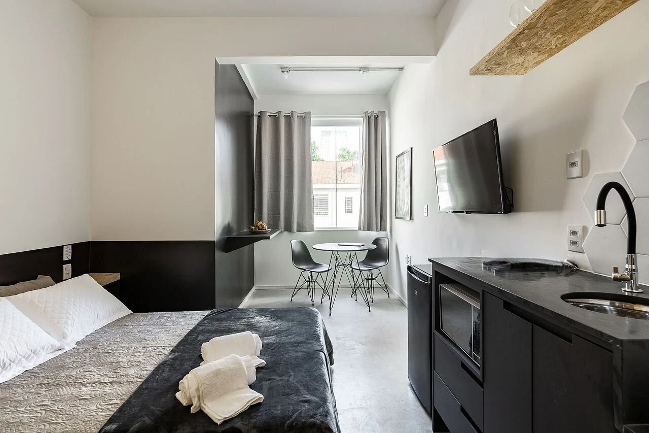 A hotel room with a bed, a desk, a chair, a TV, and a sink.