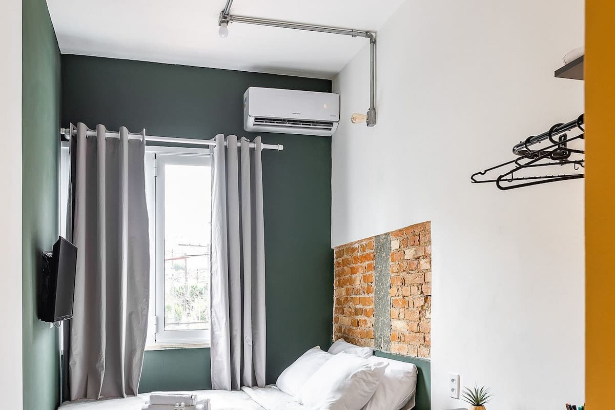 A compact bedroom with a bed, a desk, a chair, an air conditioner, and a window with curtains.