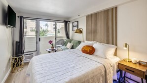 A hotel room with a bed, a bedside table with a lamp, a small table with a chair, a TV mounted on the wall, and a window with curtains. - Bright Cozy Condo Unit With Balcony (Honolulu)