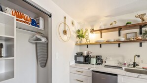 A modern kitchen with white cabinetry, a built-in oven, and a stainless steel dishwasher. There are shelves above the counter holding decorative items and a microwave. - Bright Cozy Condo Unit With Balcony (Honolulu)