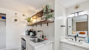 A modern kitchen with a built-in oven, a sink, and a microwave. There are wooden shelves above the counter holding decorative items and plants. - Bright Cozy Condo Unit With Balcony (Honolulu)