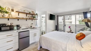 A modern kitchen with white cabinets and stainless steel appliances, a dining area with a table and chairs, and a large bed with a white comforter and decorative pillows. - Bright Cozy Condo Unit With Balcony (Honolulu)