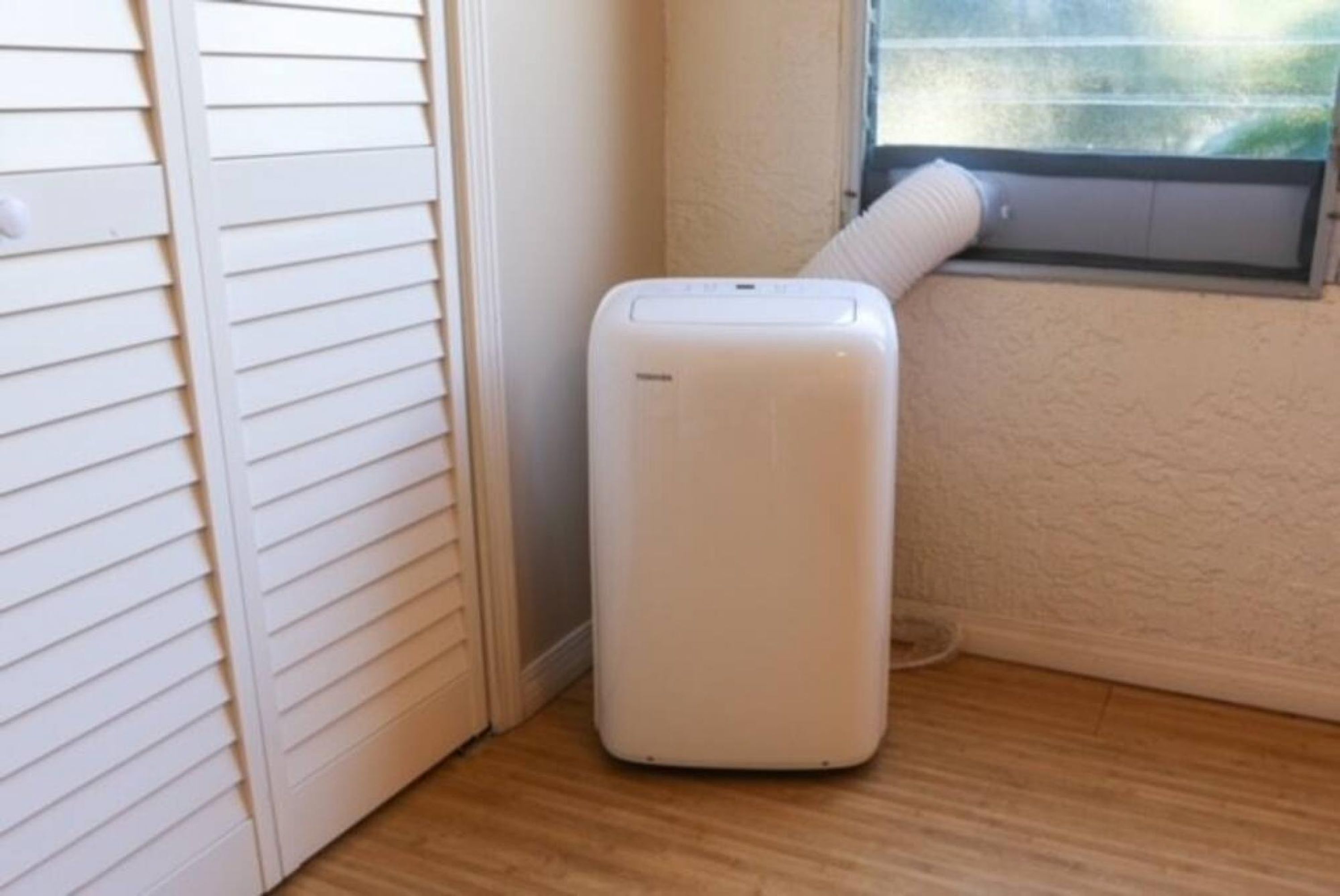 Un climatiseur portatif blanc, placé dans un coin d’une pièce dont le sol est en bois et qui possède une fenêtre.