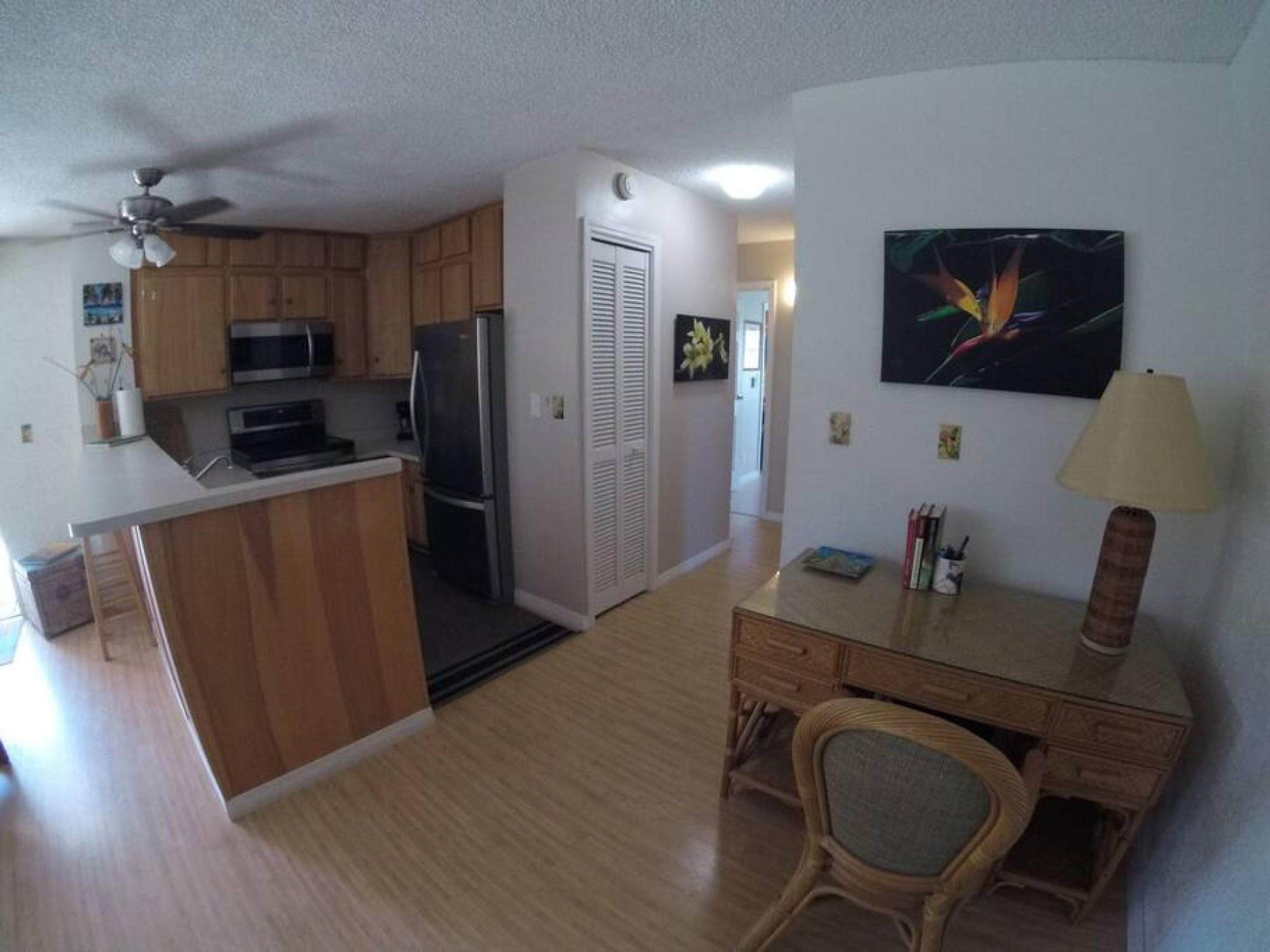Una cocina moderna con armarios de madera, microondas y refrigerador. Hay un comedor con un escritorio y una silla, y un cuadro en la pared.